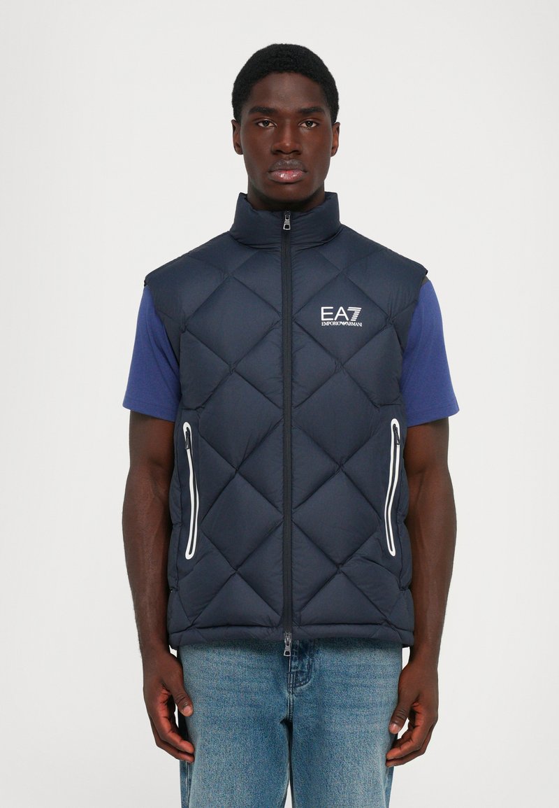 Homme portant un gilet sans manches en quilté bleu marine avec fermeture éclair à l'avant et logo EA7 Emporio Armani, sur une chemise à manches courtes bleue et un jean en denim.