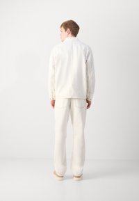 White cotton jacket with snap buttons, standing collar, and back seam detail; paired with white cotton pants and light shoes.