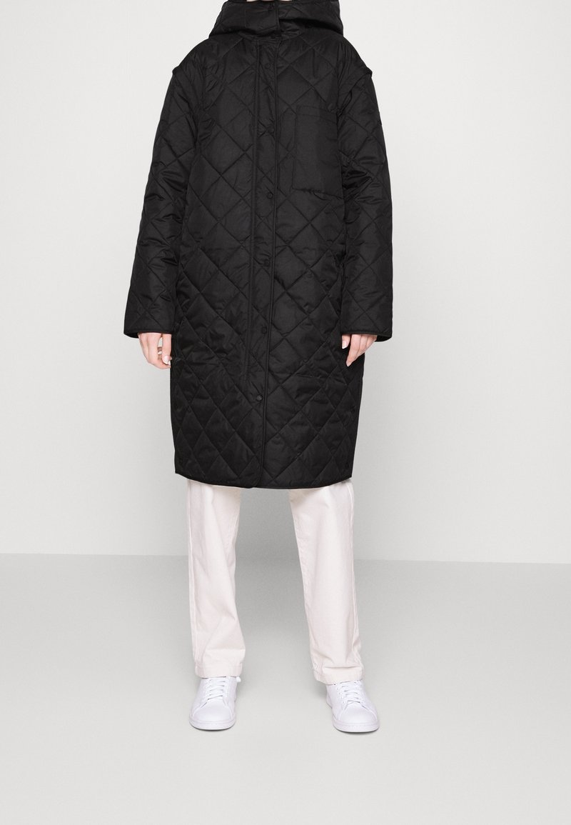 Abrigo largo negro acolchado con capucha, con cremallera frontal, grandes bolsillos parche y silueta recta, combinado con pantalones de color claro.