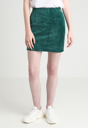 Kobieta ubrana w ciemnozieloną mini spódniczkę zamszową, białe tenisówki i biały top, stojąca na tle jasnego, jednolitego tła.