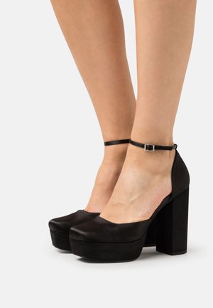 Pieds portant des escarpins à plateforme noire avec des brides à la cheville sur fond blanc.