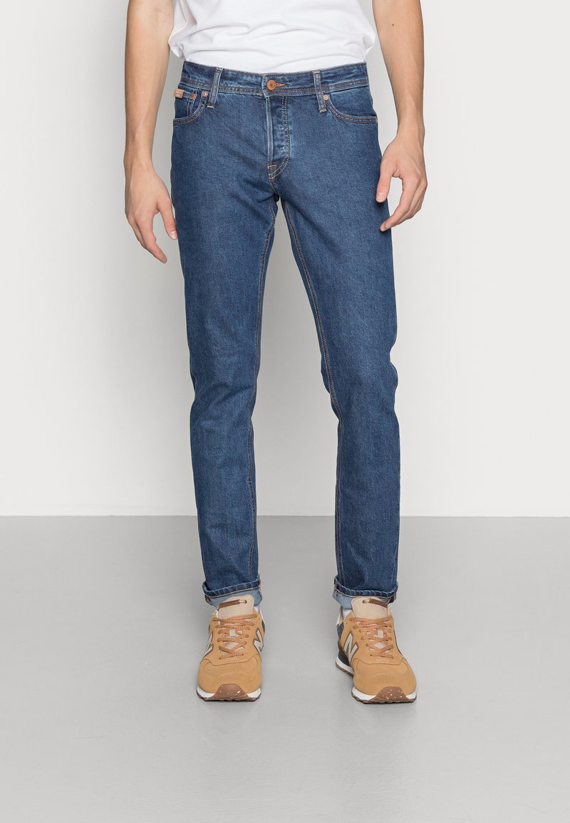 Jeans in denim blu con una vestibilità slim, cuciture a contrasto e chiusura a bottone. Abbinati a sneakers sportive color sabbia e calzini bianchi.