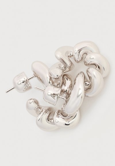 Χοντρά ασημένια σκουλαρίκια κρίκοι με στριφογυριστό, γυαλιστερό, ακανόνιστο σχέδιο και κλείσιμο με κούμπωμα καρφί σε απλό φωτεινό φόντο.