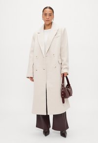 Φωτεινό μπεζ διπλόχρωμο παλτό με υφή, μεγάλα καμπυλωτά γιακά και δύο κουμπιά, συνδυασμένο με καφέ παντελόνι φαρδιάς γραμμής και μια μπορντό τσάντα.