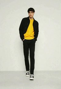 Casaco preto, suéter amarelo em malha, calças pretas e ténis com padrão em preto e branco. Modelo posa com as mãos nos bolsos.