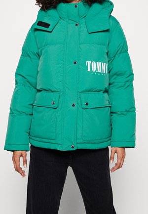 Vihreä toppatakki, jossa edessä painonapit, kaksi läppätaskua ja valkoinen "TOMMY JEANS" -logo rinnassa, mustia housuja käyttävän henkilön päällä.