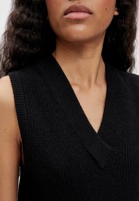 Maglione senza maniche nero con scollatura a V; tessuto a coste testurizzato; primo piano del collo e della parte superiore.