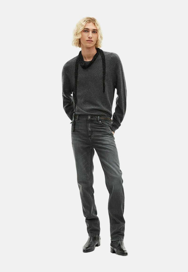 Grauer Pullover mit lockerer Passform und einem drapierten schwarzen Band. Dunkelgraue Jeans mit geradem Schnitt. Schwarze Schuhe vervollständigen das Outfit.