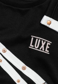 Tessuto nero con rifiniture bianche e bottoni a pressione in rame, con la parola "LUXE" in lettere maiuscole bianche.
