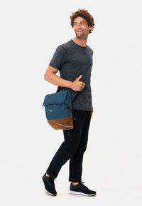 Borsa a tracolla blu con dettagli marroni, in materiale tessile, con tracolla regolabile e particolari riflettenti. Modello che indossa una maglietta scura e pantaloni neri.