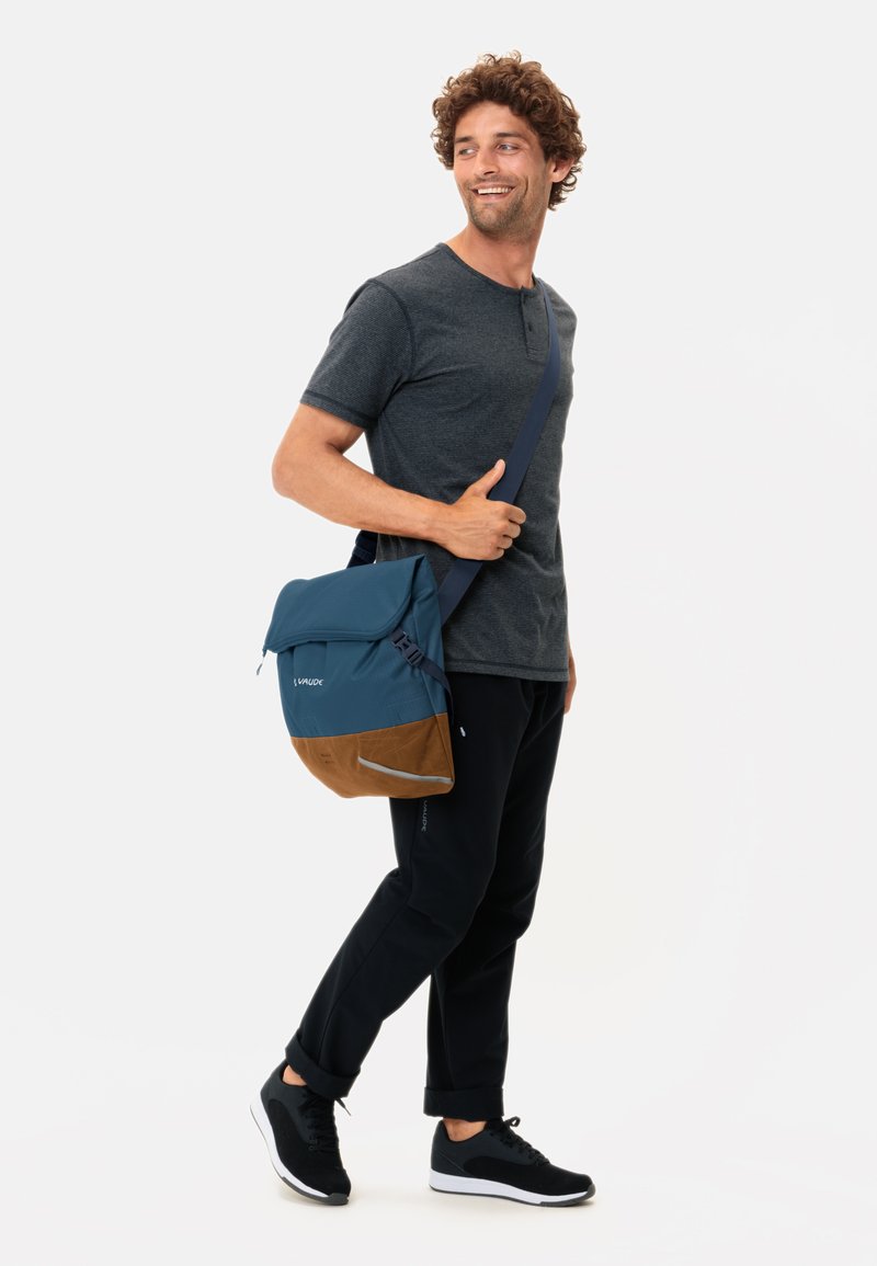 Borsa a tracolla blu con dettagli marroni, in materiale tessile, con tracolla regolabile e particolari riflettenti. Modello che indossa una maglietta scura e pantaloni neri.