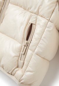 Sezione di giacca a vento beige con tessuto lucido e trapuntato, caratterizzata da una cerniera con linguetta coordinata e dettagli di cucitura visibili.