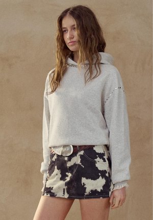 Jonge vrouw met lang golvend haar, gekleed in een lichtgrijze hoodie met studs en een zwart-wit minirok met koeienprint en een grote gesp.