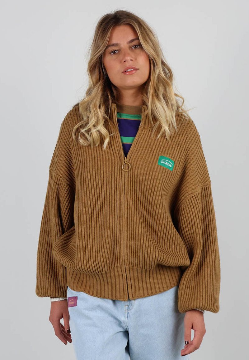 Pull en maille côtelée marron à fermeture éclair avec une fermeture à glissière en métal rond et une étiquette verte ; doté de manches larges et d'une coupe décontractée. Porté par-dessus une chemise rayée.