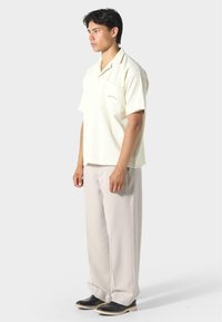 Světle žlutá, krátkozásadová textilní košile s kapsou, v kombinaci s volnými, světle šedými kalhotami a černými botami. Profil postavy z boku.