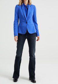 Blå blazer med slag och en knapp, parat med mörkblå jeans, bärs med en matchande blå skjorta under.