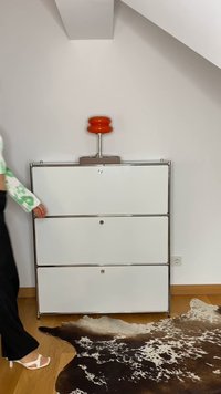 Hvit tre-skuffers skap med kromdetaljer, toppet med en oransje lampe. Et teksturert svart-hvitt cowhide-teppe er delvis synlig.