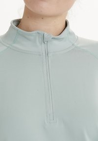 Ljusblå zip-up atletisk topp i elastisk tyg, med hög krage och kontrasterande sömmar. Slät textur, åtsittande design.