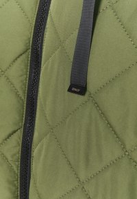 Tissu matelassé vert avec un motif en diamond, doté d'une fermeture éclair noire et d'une sangle avec l'étiquette "ONLY" près du haut. Texture lisse dans l'ensemble.