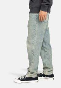 Vaqueros de mezclilla de color azul claro y deslavado, con corte recto, que cuentan con un bolsillo trasero y una textura sutil. Combinados con zapatillas de lona negras.