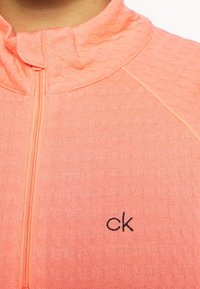 Haut zippé corail vif en tissu texturé. Il présente un col montant et un petit logo noir "ck" sur le côté gauche.