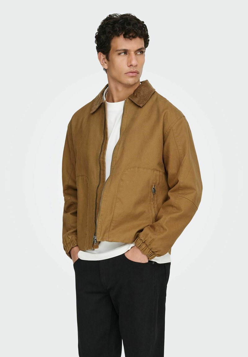 Moški s kodrasto temno frizuro, oblečen v rjav zadrgnjen jopič čez belo srajco in črne hlače, z rokami v žepih, gleda na stran.