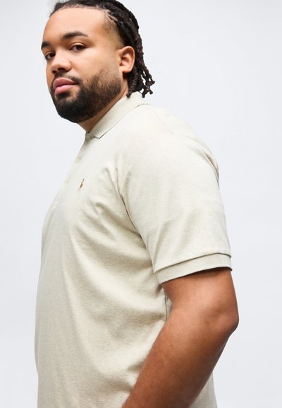 Άντρας με γενειάδα και πλεξούδες, φορώντας μπεζ πόλο μπλουζάκι, σε πλάγια όψη με φόντο ένα απλό λευκό τοίχο.