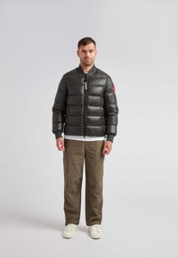 Giacca a vento nera con chiusura a zip, maniche corte, polsini a coste e un accento rosso sul braccio sinistro. Abbinata a pantaloni beige e scarpe da ginnastica bianche.