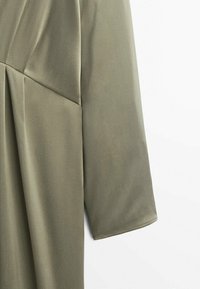Gros plan d'une manche longue en tissu vert olive clair avec un design de couture près de l'épaule sur fond blanc.