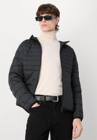 Svart puffjacka med dragkedja fram, figursydd design och ribbade ärmslut; bärs över en ljusbeige polotröja och svarta byxor med bälte.