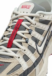 Nike sneaker featuring a gray mesh upper, beige suede accents, white laces, and a red pull tab with a black swoosh logo.