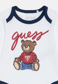 Biely bavlnený overal s námorníckym lemovaním, na ktorom je kartónový medveď v červenom svetri a modrých džínsach, s potlačou "Guess" v tučnom písme.