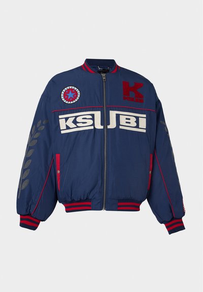 Blouson bomber bleu marine avec des rayures rouges et blanches, fermeture éclair à l'avant, patches rouges, texte "KSUBI" et motifs de feuilles sur les manches.