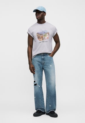 Grå grafisk t-skjorte med oppbrettede ermer; blå slitte vidbukser; svarte sko; lyseblå caps med tekst.