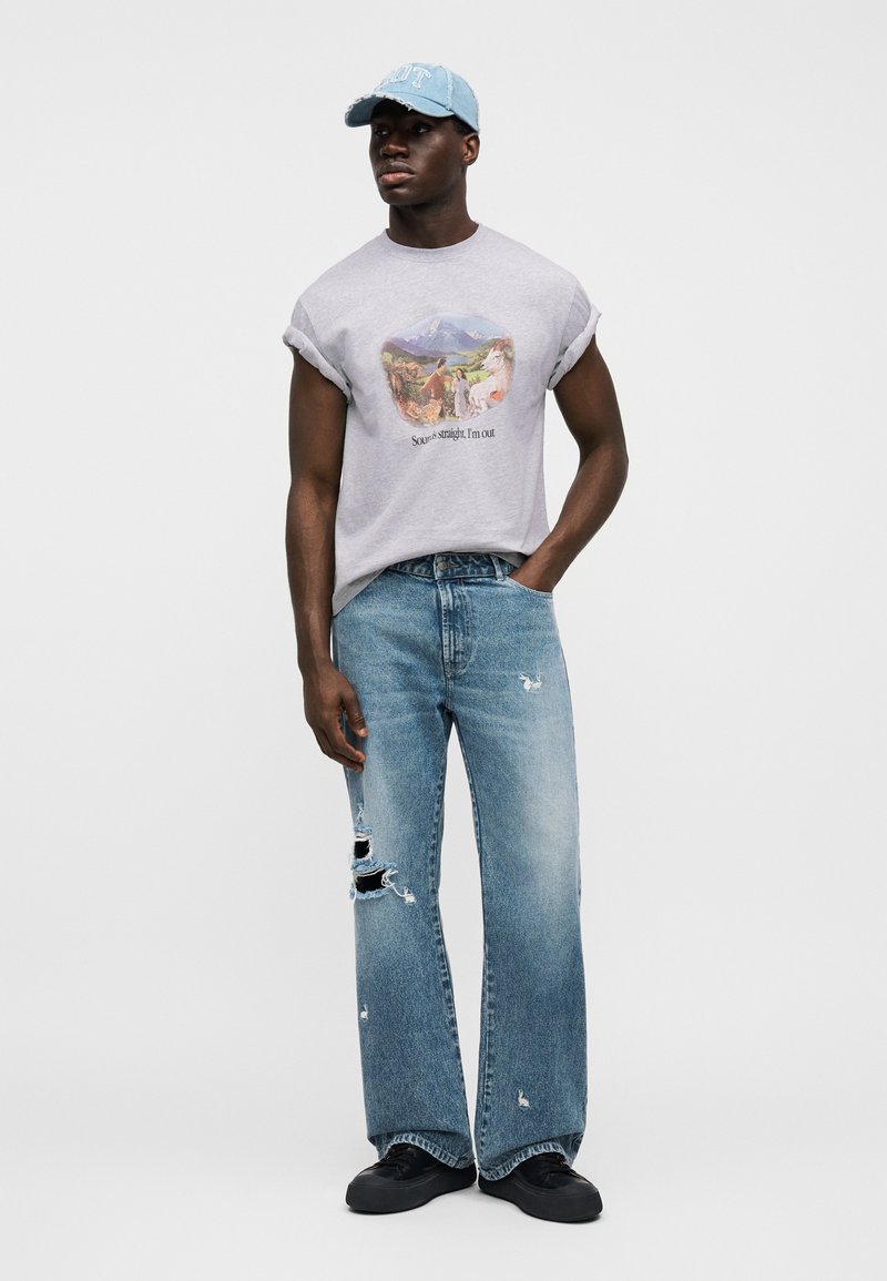 Sivé grafické tričko s vyhrnutými rukávmi; modré roztrhané džínsové nohavice širokého strihu; čierne topánky; svetlomodrá čiapka s textom.