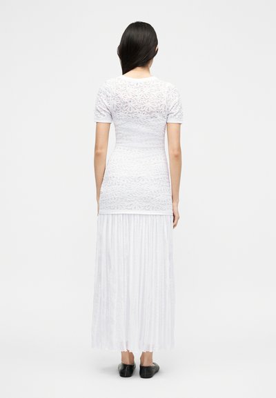 Žena s dlouhými tmavými vlasy, která má na sobě krátký bílý krajkový top s krátkým rukávem a dlouhou plisovanou bílou sukni, stojící zády k divákovi na jednobarevném pozadí.