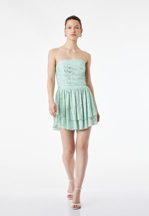 Donna che indossa un mini abito verde chiaro senza spalline con macchie nere e gonna a strati, abbinato a tacchi bianchi con cinturino alla caviglia, in piedi su sfondo bianco.