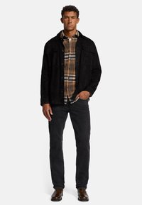 Veste en daim noire sur une chemise à carreaux brune, avec deux poches poitrine ; associée à un pantalon texturé noir et des bottes en cuir marron.