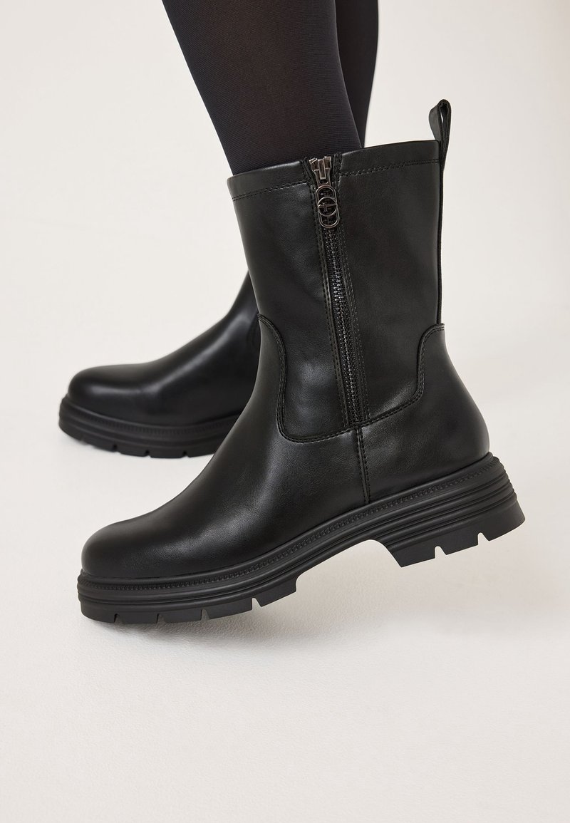 Bottes en cuir noir mi-mollet avec fermeture éclair latérale et semelles épaisses portées avec des collants noirs sur fond pâle.