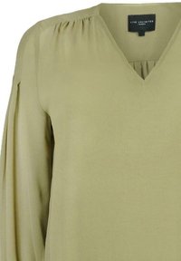 Olivově zelená halenka s výstřihem do V a jemnou texturou; má na ramenou nabrání a dlouhé rukávy. Na štítku je nápis "LIVE UNLIMITED LONDON."