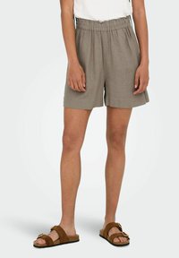 Pantaloni corti leggeri di colore khaki con vita elastica e tasche laterali. Indossati con sandali marroni con un dettaglio a fibbia.