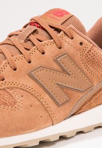 Gros plan d'une basket New Balance en daim couleur sable avec logo brodé, lacets épais et semelle texturée sur fond blanc.