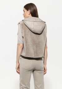 Frau in beigem Kurzarmoberteil, taupefarbenem Kunstpelz-Weste mit gepolstertem Kapuzen, und beigen Hosen, von hinten auf einem weißen Hintergrund gezeigt.