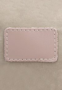 Beige kangas, jossa keskellä ommeltu sileä, vaaleanpunainen nahkainen etiketti. Etiketissä on hillitty ommeltu reunus.