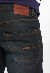 Mørke denimjeans med nært billede af bagsiden, der har orange syninger, to baglommer og et brunt læder ROC-A-WEAR skilt.