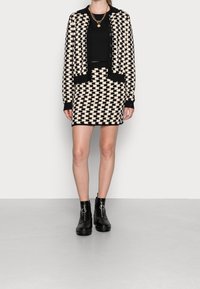 Un cardigan à motif noir et crème porté sur un top noir court, associé à une jupe assortie avec le même motif et des bottes de cheville noires zippées.