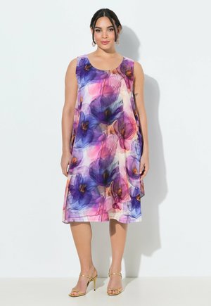 Donna con vestito senza maniche con grandi motivi floreali viola e rosa e sandali dorati con tacco alto, in piedi contro un muro bianco.