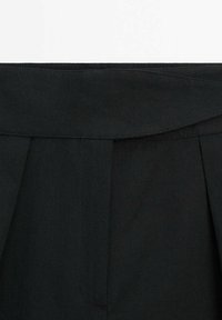 Zwarte op maat gemaakte broeken hebben een zachte stofstructuur, een brede tailleband en scherpe plooien, zonder zichtbare hardware of accenten.