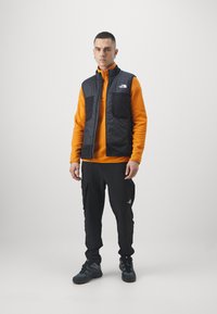 Black insulated vest over an orange zip-up fleece, paired with black cargo pants and gray hiking shoes. Features include multiple pockets and logos.