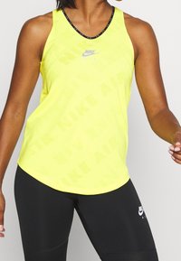 Top sin mangas amarillo de Nike con un patrón de logo sutil, diseño de espalda nadadora y escote negro en contraste, combinado con leggings negros que presentan un pequeño logo.